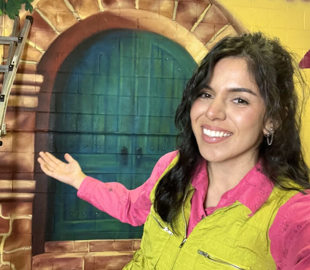 Artist Camila Ibarra presents her colorful mural, smiling and gesturing towards her artwork, at a Paint and Sip event.
