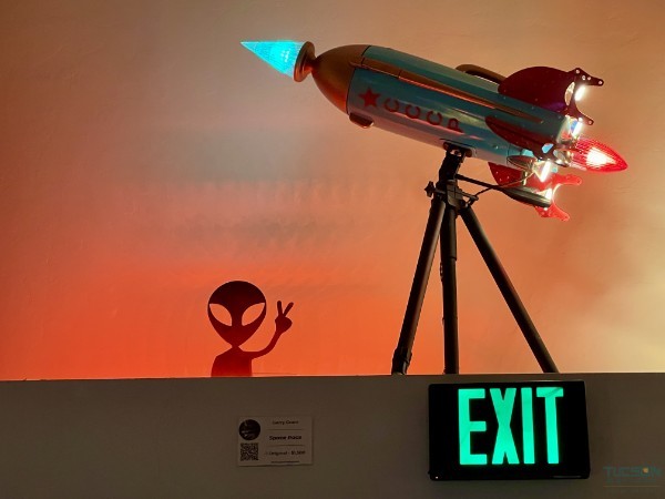 Space Race by Gerry Grant, an illuminated rocket ship sculpture displayed above an exit sign with glowing lights and an alien silhouette.